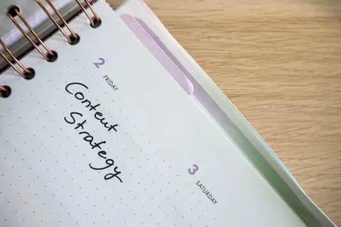 Draufsicht auf ein aufgeschlagenes Notizbuch mit Tagesplaner, in dem handschriftlich &bdquo;Content Strategy&ldquo; steht, auf einem Holzschreibtisch.