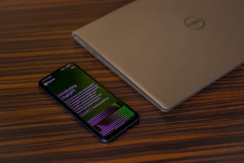 Ein Smartphone mit der ChatGPT-App neben einem zugeklappten Laptop auf einem Holztisch.