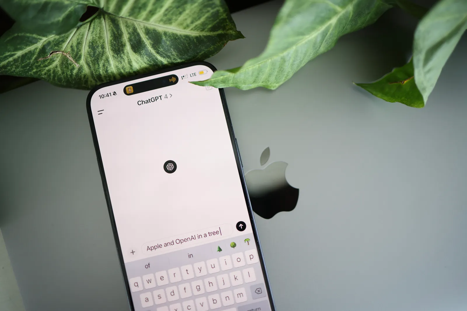 A smartphone shows a ChatGPT interface placed on an Apple laptop in a leafy environment.