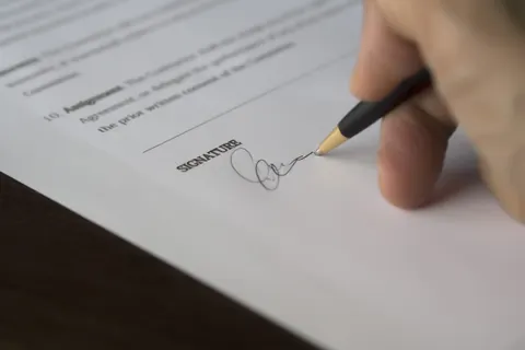 Eine Hand unterschreibt auf einem Holzschreibtisch mit einem Stift einen offiziellen Vertrag.