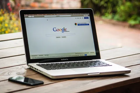 Ein MacBook Pro mit ge&ouml;ffneter Google-Suche auf einem Holztisch im Freien, neben einem Smartphone.
