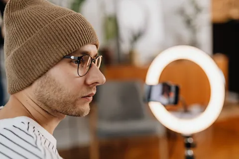 Ein Mann mit Brille und Strickm&uuml;tze nimmt zu Hause mit Smartphone und Ringlicht ein Video auf.