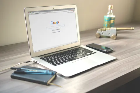 Aufger&auml;umter Arbeitsplatz auf einem Holzschreibtisch: Laptop mit ge&ouml;ffneter Google-Suche, Smartphone und Notizbuch.