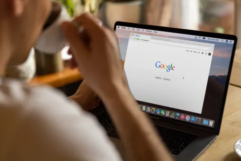 Nahaufnahme einer erwachsenen Person, die drinnen Kaffee trinkt und auf einem Laptop Google nutzt.