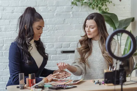 Junge, gut gelaunte Bloggerinnen unterschiedlicher Herkunft testen am Tisch Make-up-Produkte und nehmen mit einem Smartphone auf einem Stativ mit Ringlicht einen Vlog auf