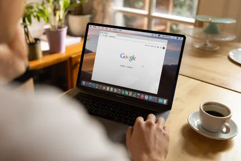 Eine erwachsene Person sitzt drinnen mit Kaffee an einem Holztisch und nutzt auf dem Laptop Google.