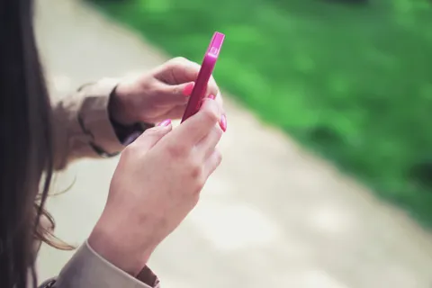 Frau h&auml;lt in entspannter Umgebung im Freien ein rosa Smartphone mit beiden H&auml;nden.