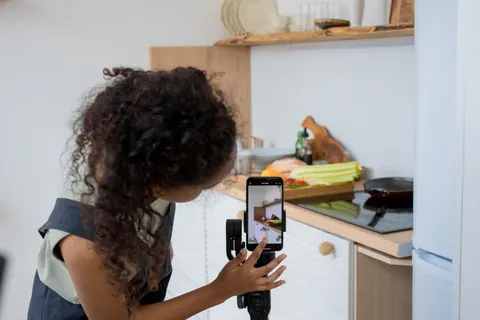 Eine Frau mit lockigem Haar filmt in einer modernen K&uuml;che mit einem Smartphone-Stabilisator ein Kochvideo.
